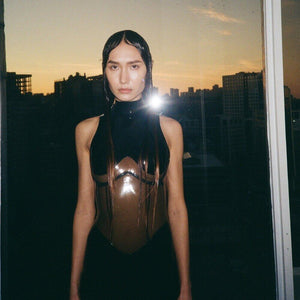 Body en latex noir ANOESES avec panneaux transparents et opaques, porté par un modèle debout devant une fenêtre avec vue urbaine au coucher du soleil.