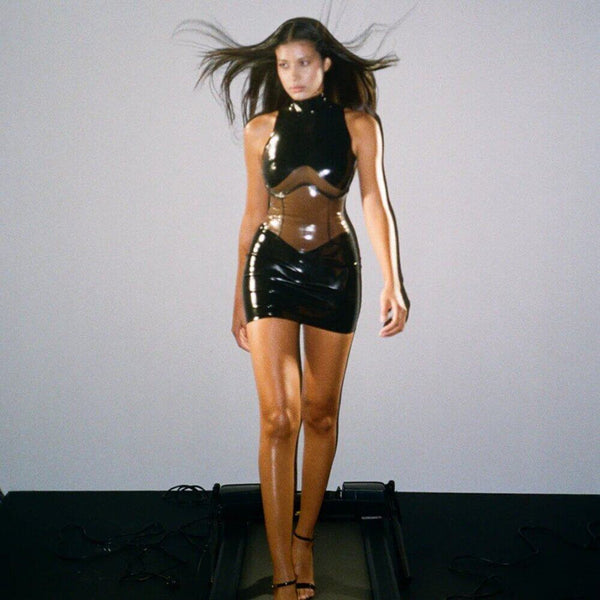 Body en latex noir ANOESES avec col montant et empiècements transparents, porté par un mannequin assis sur un fauteuil en cuir noir, vue légèrement plongeante.