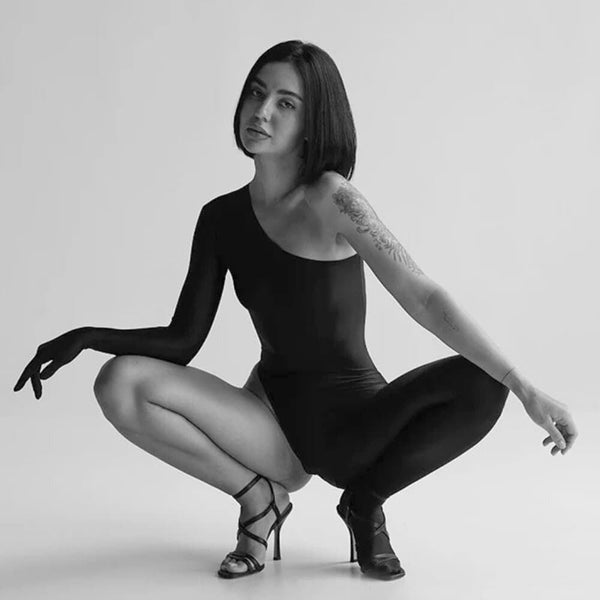 Catsuit DEXTRA ANOESES porté par un modèle en pose accroupie, pièce asymétrique noire seconde peau, sélection Brigade Mondaine Paris.