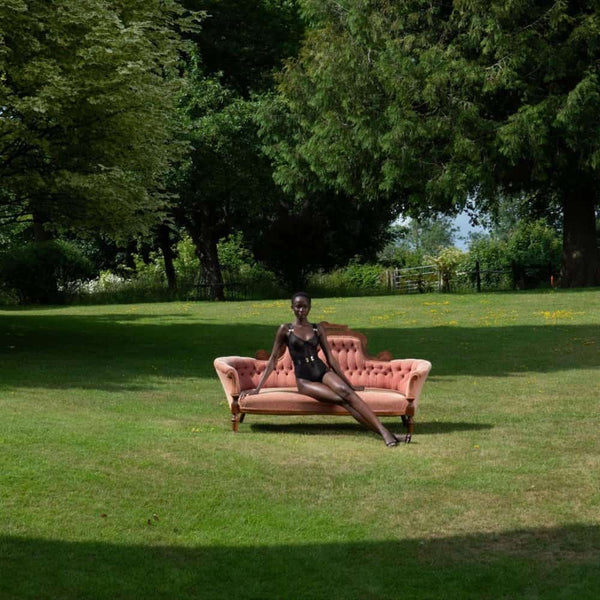 Mannequin assis sur un canapé ancien rose poudré posé sur une pelouse, portant un body noir BORDELLE, dans un vaste jardin arboré.