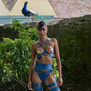 Mannequin portant un ensemble BORDELLE bleu composé de sangles elastiques, ornements dorés en forme de coquille et bas taille haute, photographié dans un jardin avec un paon posé sur un muret en pierre à l’arrière-plan.