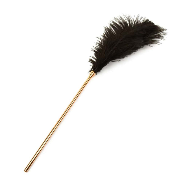 Set BDSM en cuir noir composé de liens ajustables avec anneaux dorés, d’un bandeau en tissu satiné et d’une plume noire montée sur manche doré, présenté sur fond blanc dans une esthétique épurée.