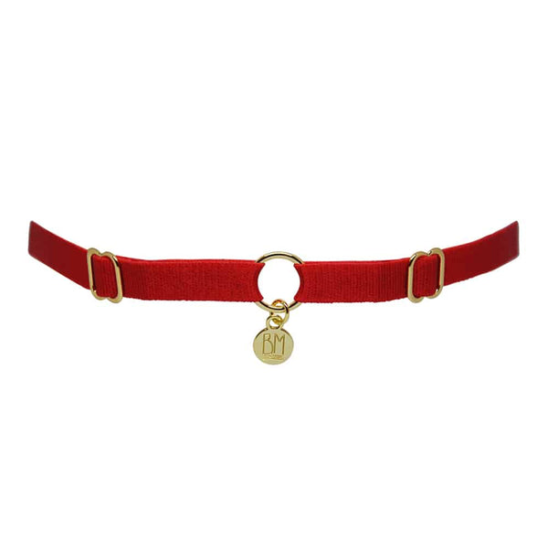 Ici vous pouvez voir le BRIGADE MONDAINE GIFT WRAP RED. Ce collier est fait d’une bande rouge. 2 détails pour régler les bandes se trouvent à droite et à gauche. Au milieu, la bande est séparée par un anneau plaqué or avec un pendentif où il est inscrit « BM ».