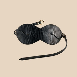 Masque BDSM en cuir noir texturé à la forme enveloppante, avec attaches réglables et bouclerie dorée, présenté sur fond noir dans une mise en scène élégante et minimaliste.