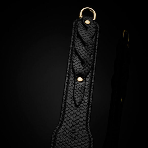 Modèle portant un body noir transparent et tenant un paddle BDSM en cuir noir à finition texturée, photographiée sur fond blanc dans une mise en scène épurée et élégante.