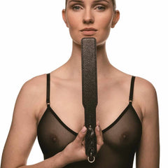 Modèle portant un body noir transparent et tenant un paddle BDSM en cuir noir à finition texturée, photographiée sur fond blanc dans une mise en scène épurée et élégante.