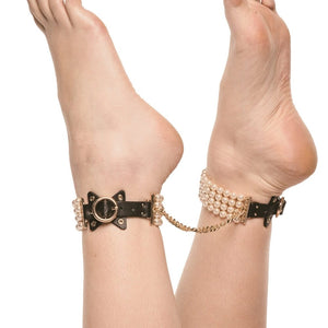 Entraves de chevilles en cuir noir ornées de perles et reliées par une chaîne dorée, portées sur modèle, proposées par Brigade Mondaine Paris.