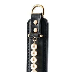 Paddle BDSM en cuir noir avec finition lisse et rangée de perles décoratives, tenu par un modèle en studio, accessoire sélectionné par Brigade Mondaine Paris.