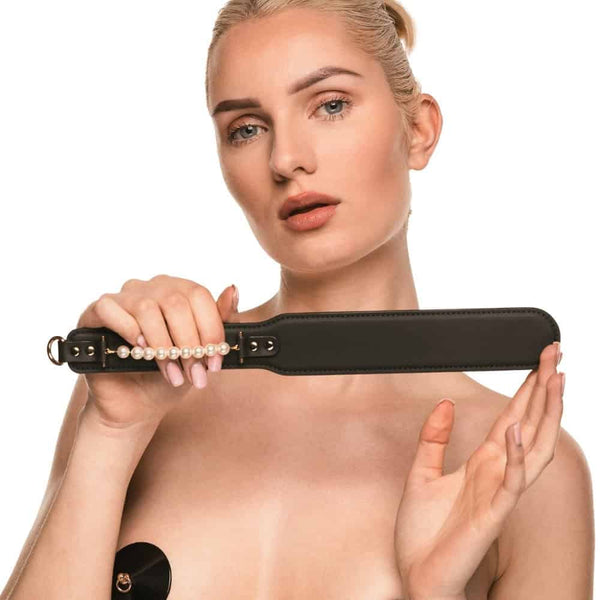 Paddle BDSM en cuir noir avec finition lisse et rangée de perles décoratives, tenu par un modèle en studio, accessoire sélectionné par Brigade Mondaine Paris.