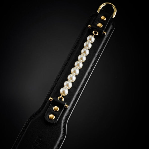 Paddle BDSM en cuir noir avec finition lisse et rangée de perles décoratives, tenu par un modèle en studio, accessoire sélectionné par Brigade Mondaine Paris.