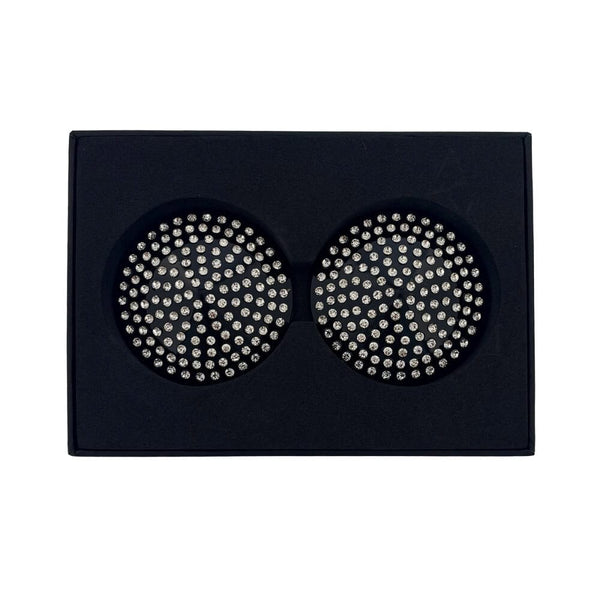 Paire de cache-tétons noirs circulaires ornés de strass brillants, présentés dans un écrin noir sur fond blanc, avec une esthétique élégante et minimaliste.