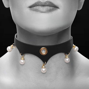 Gros plan d’un collier BDSM en cuir noir porté au cou, orné d’anneaux dorés et de perles blanches, sur fond sombre, dans une esthétique élégante et minimaliste.