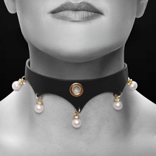 Gros plan d’un collier BDSM en cuir noir porté au cou, orné d’anneaux dorés et de perles blanches, sur fond sombre, dans une esthétique élégante et minimaliste.