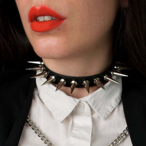 Gros plan sur le choker 86137, porté par une femme avec rouge à lèvres rouge, chemise blanche et blazer noir orné de deux chaînes argentées. Le choker est orné de spikes métalliques de 26 mm et d'une largeur de 1,5 cm, avec un ajustement réglable à l'arrière.