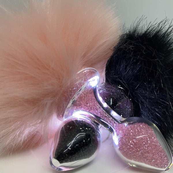 Image de deux plugs disposés en superposition, l'une de couleur rose et l'autre noire, remplies de billes éclatantes de leurs teintes respectives, chacune ornée d'une queue de lapin suspendue.