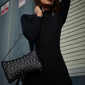 Sac en cuir noir au design sophistiqué, orné de chaînes en argent et d’un pompon élégant, avec une hanse supplémentaire. Mis en scène dans une photo haut de gamme, porté par une femme en robe noire chic pour un look luxueux et raffiné.