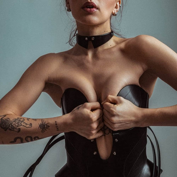 Mannequin portant le corset Daelos en cuir noir RE:STRICT chez Brigade Mondaine Paris, décolleté plongeant structuré, sangles ajustables, porté avec accessoires assortis sur fond rouge.
