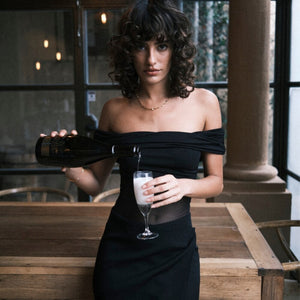 Body noir Undress Code Libra à épaules dégagées, porté par un modèle tenant une coupe de champagne, sélection Brigade Mondaine Paris.