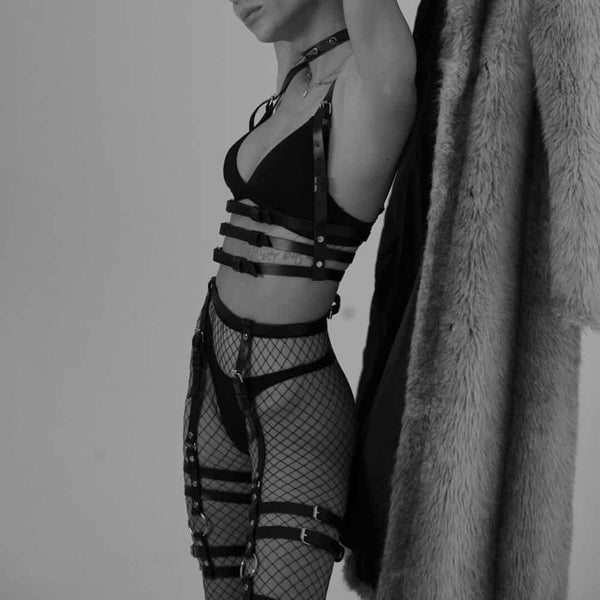 Dans une ambiance en noir et blanc, une femme porte le Harnais CLASSIC 3 en cuir – ST. VENDETTA et les jarretières Gorgona en cuir, mettant en valeur sa silhouette. Elle tient son manteau en fourrure derrière elle, accentuant son allure audacieuse. Sous le manteau, elle porte un collant résille et un soutien-gorge noir, apportant une touche de sensualité supplémentaire et renforçant l’intensité de son style raffiné.