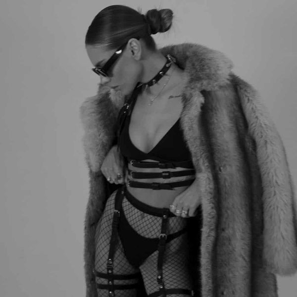 Femme en noir et blanc portant un harnais en cuir naturel fait à la main, avec trois lanières sous le buste et deux lanières montant vers le cou, créant un effet de soutien-gorge. Elle porte également un soutien-gorge noir, et le cuir est mis en valeur par la lumière, accentuant la texture et le design sophistiqué.