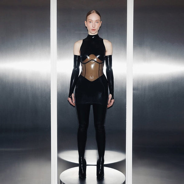 Combinaison en latex noir ANOESES avec empiècement transparent au buste, manches longues et col montant, portée par un modèle debout dans un décor minimaliste.
