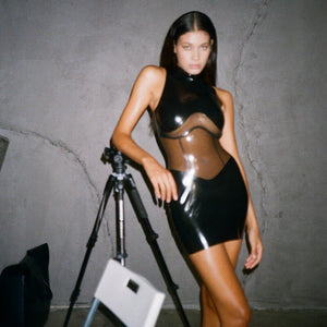 Body en latex noir ANOESES avec col montant et empiècements transparents, porté par un mannequin assis sur un fauteuil en cuir noir, vue légèrement plongeante.