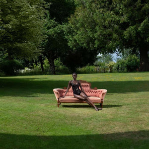 Mannequin assis sur un canapé ancien rose poudré posé sur une pelouse, portant un body noir BORDELLE, dans un vaste jardin arboré.