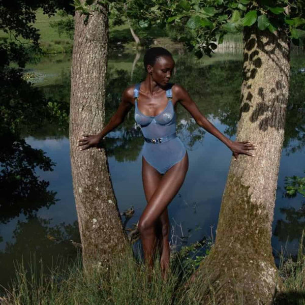 Mannequin debout entre deux arbres, portant un body BORDELLE bleu aux lignes structurées, au bord d’un étang, dans un décor naturel verdoyant.