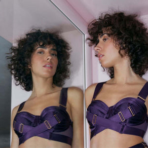Une femme debout se regardant dans le miroir et portant le Soutien-Gorge Bustier en violet avec de larges bretelles amovibles et des ajustements dorés.