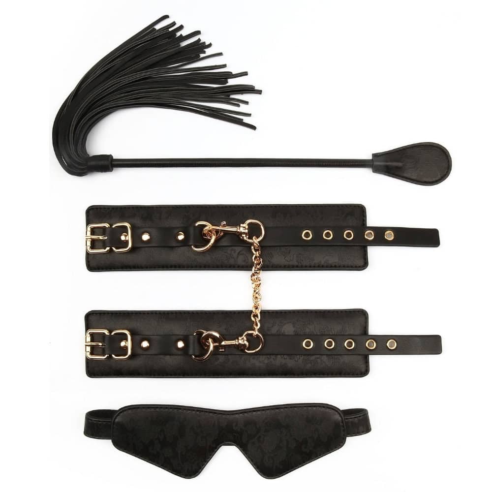Set BDSM en cuir noir comprenant une paire de menottes reliées par chaîne dorée, un masque occultant et une cravache assortie, présenté sur fond blanc dans une mise en scène élégante.