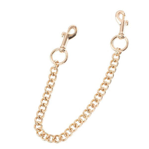 Paire de menottes de chevilles en cuir noir reliées par une chaîne dorée, avec boucles métalliques ajustables et finitions soignées, présentées sur fond blanc dans une esthétique épurée.