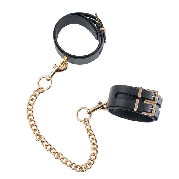 Paire de menottes de chevilles en cuir noir reliées par une chaîne dorée, avec boucles métalliques ajustables et finitions soignées, présentées sur fond blanc dans une esthétique épurée.