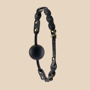 Bâillon boule BDSM composé d’une boule noire mate et de sangles en cuir noir texturé avec rivets et bouclerie dorée, présenté sur fond sombre dans une mise en scène élégante.