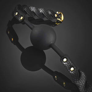 Bâillon boule BDSM composé d’une boule noire mate et de sangles en cuir noir texturé avec rivets et bouclerie dorée, présenté sur fond sombre dans une mise en scène élégante.