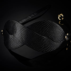 Masque BDSM en cuir noir texturé à la forme enveloppante, avec attaches réglables et bouclerie dorée, présenté sur fond noir dans une mise en scène élégante et minimaliste.