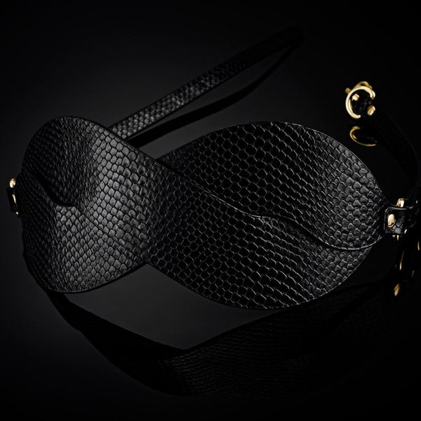 Masque BDSM en cuir noir texturé à la forme enveloppante, avec attaches réglables et bouclerie dorée, présenté sur fond noir dans une mise en scène élégante et minimaliste.