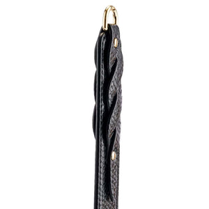 Modèle portant un body noir transparent et tenant un paddle BDSM en cuir noir à finition texturée, photographiée sur fond blanc dans une mise en scène épurée et élégante.