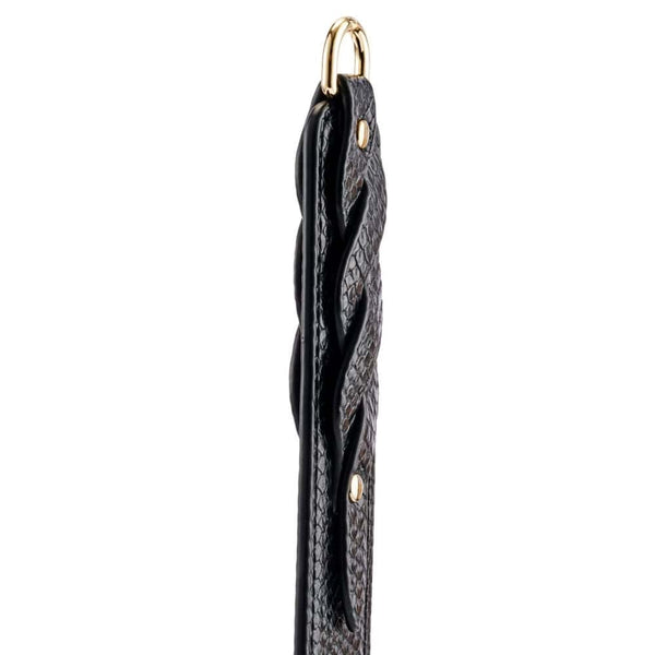 Modèle portant un body noir transparent et tenant un paddle BDSM en cuir noir à finition texturée, photographiée sur fond blanc dans une mise en scène épurée et élégante.