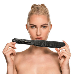 Modèle portant un body noir transparent et tenant un paddle BDSM en cuir noir à finition texturée, photographiée sur fond blanc dans une mise en scène épurée et élégante.