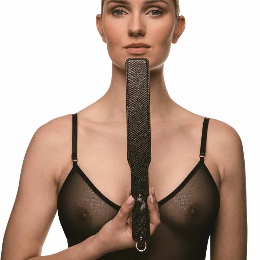 Modèle portant un body noir transparent et tenant un paddle BDSM en cuir noir à finition texturée, photographiée sur fond blanc dans une mise en scène épurée et élégante.