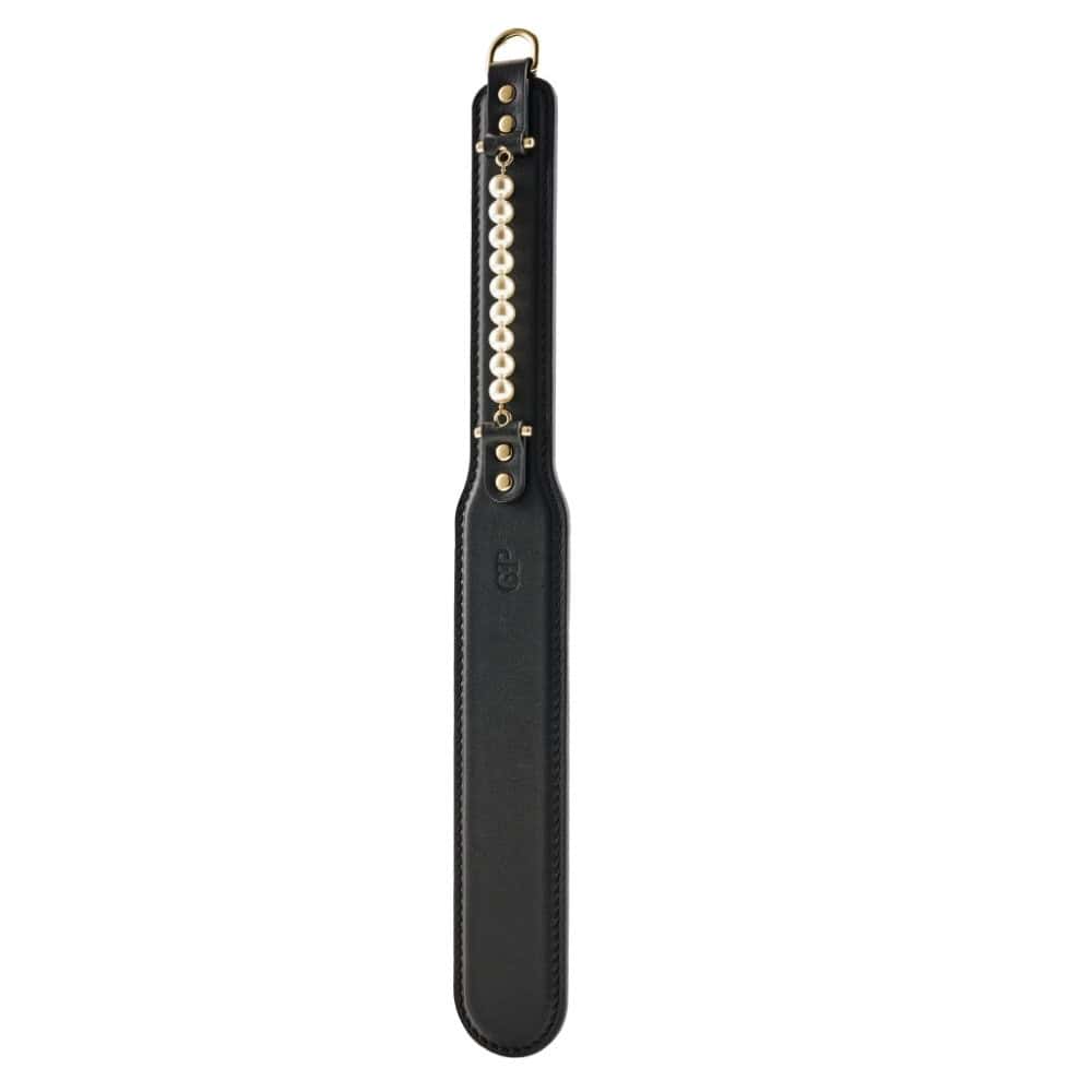 Paddle BDSM en cuir noir avec finition lisse et rangée de perles décoratives, tenu par un modèle en studio, accessoire sélectionné par Brigade Mondaine Paris.