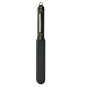 Paddle BDSM en cuir noir avec finition lisse et rangée de perles décoratives, tenu par un modèle en studio, accessoire sélectionné par Brigade Mondaine Paris.