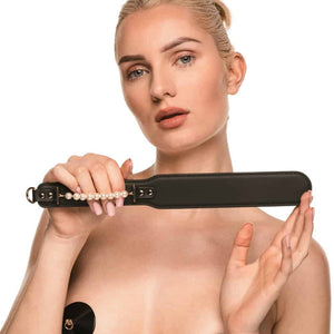 Paddle BDSM en cuir noir avec finition lisse et rangée de perles décoratives, tenu par un modèle en studio, accessoire sélectionné par Brigade Mondaine Paris.