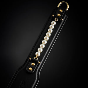 Paddle BDSM en cuir noir avec finition lisse et rangée de perles décoratives, tenu par un modèle en studio, accessoire sélectionné par Brigade Mondaine Paris.