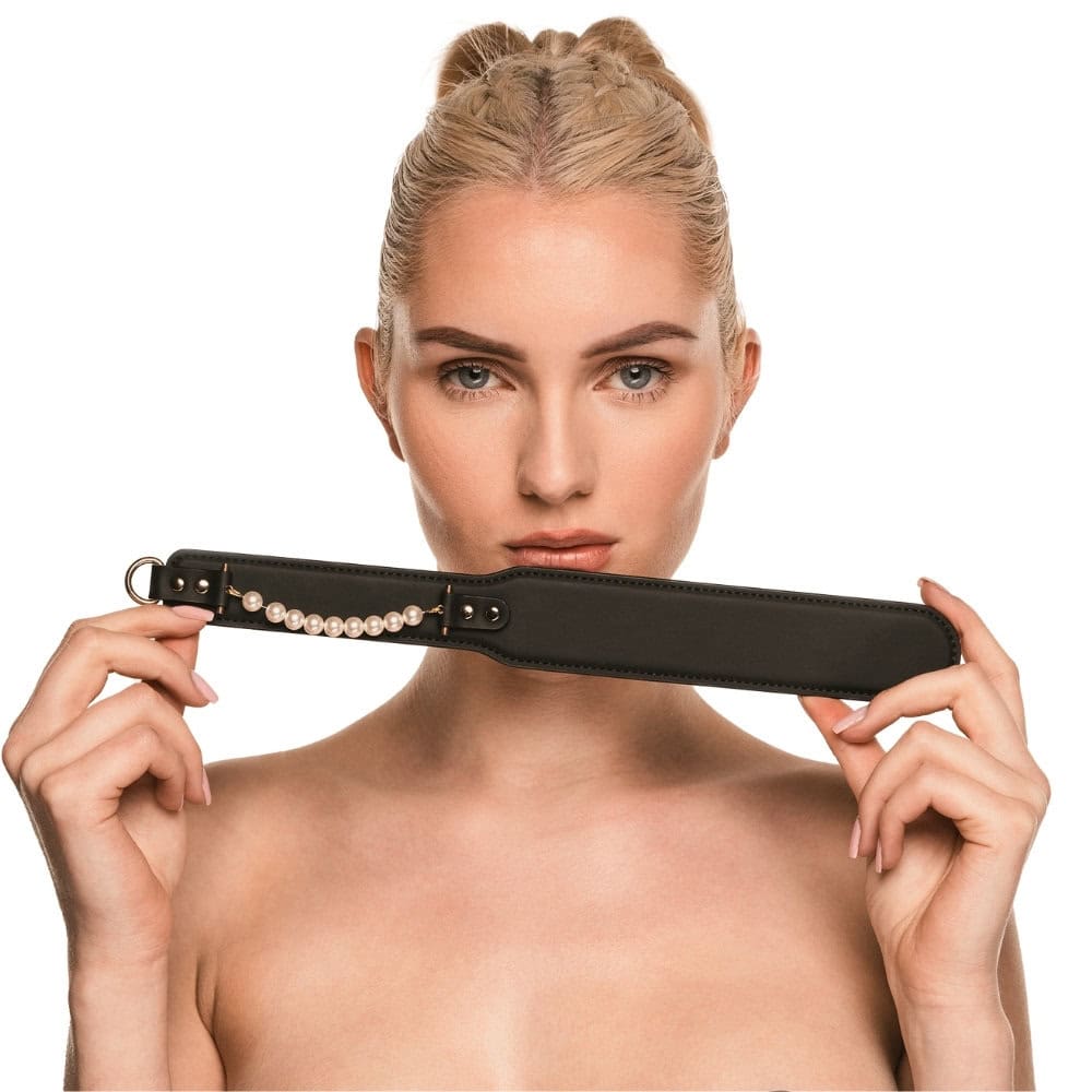 Paddle BDSM en cuir noir avec finition lisse et rangée de perles décoratives, tenu par un modèle en studio, accessoire sélectionné par Brigade Mondaine Paris.