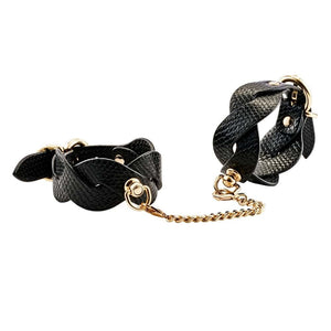 Chevilles d’un modèle portant des menottes BDSM noires texturées reliées par une chaîne dorée, accessoire de contrainte sélectionné par Brigade Mondaine Paris.