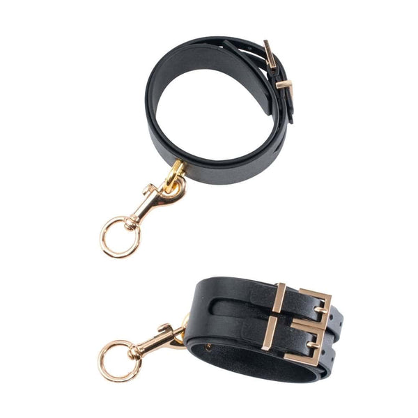 Menottes de chevilles BDSM en cuir noir avec boucle métallique et chaîne dorée, portées aux chevilles avec escarpins noirs à talons, photographiées sur fond gris.