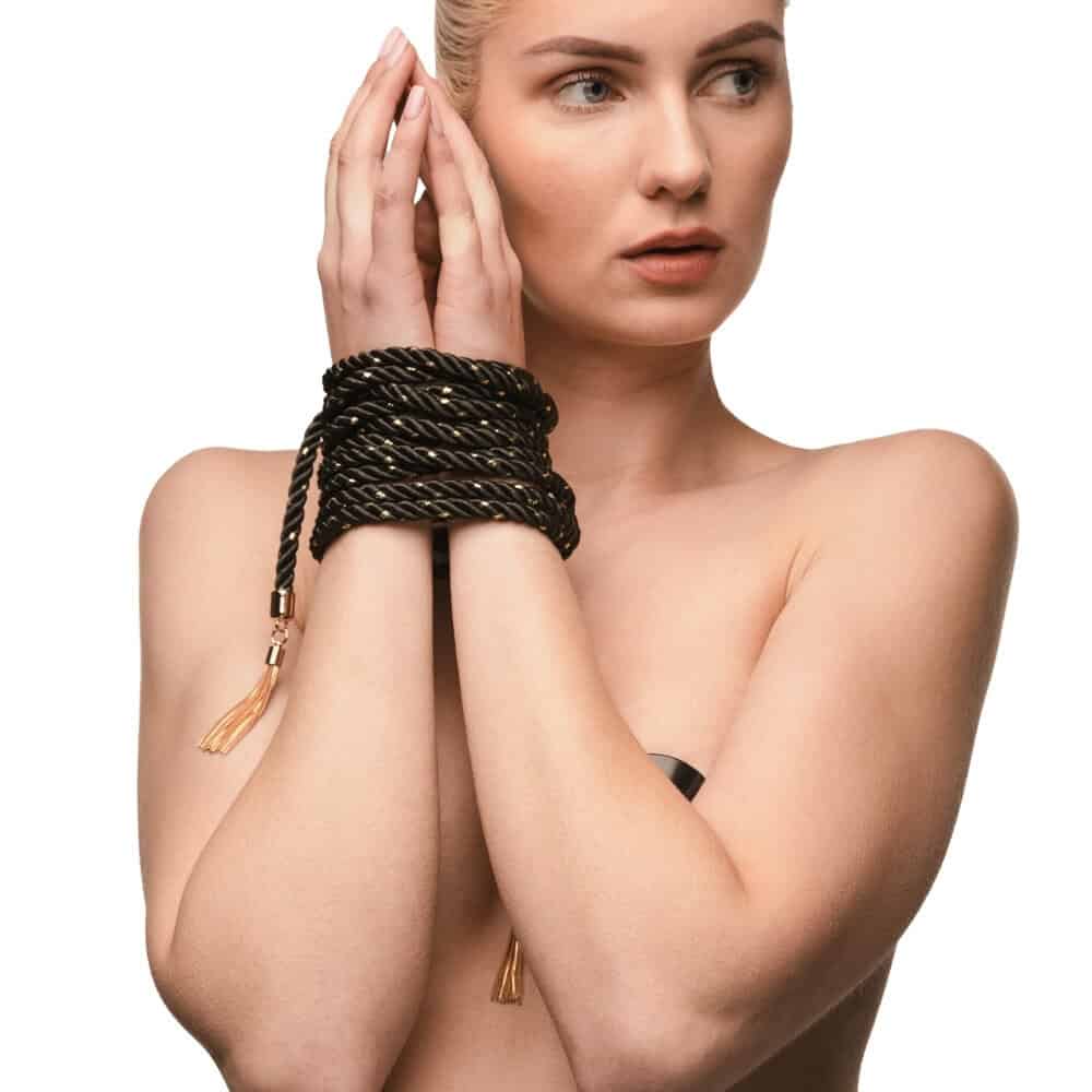 Corde BDSM noire torsadée avec incrustations dorées, fermée par une lanière en cuir et une boucle métallique, dotée de finitions dorées et pompons, présentée sur fond noir.