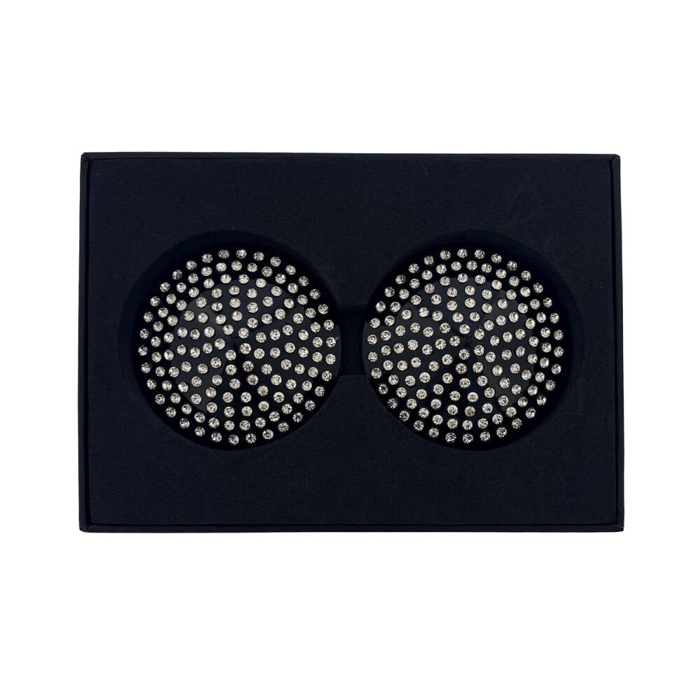 Paire de cache-tétons noirs circulaires ornés de strass brillants, présentés dans un écrin noir sur fond blanc, avec une esthétique élégante et minimaliste.