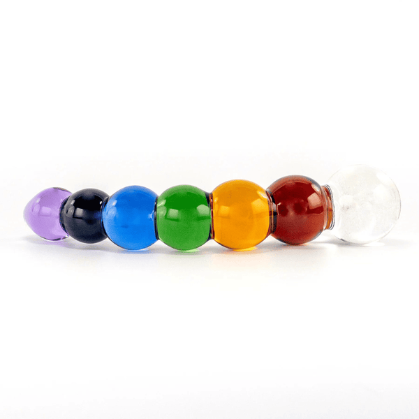 Photographie d’un dildo sur fond blanc, il représente un arc-en-ciel, il est coloré de blanc, rouge, jaune, vert, bleu, marine, violet, un festival de confettis et de couleurs